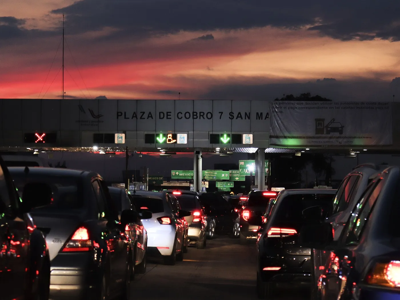 Plaza de cobro 7 de Caminos y Puentes Federales al atardecer, con filas de autos frente a las casetas, los carriles IAVE señalados y una lona de Comunicaciones y CAPUFE.