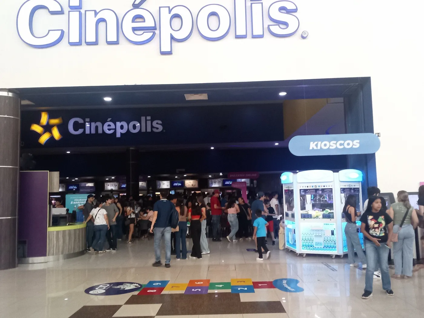 Entrada de Cinépolis en Gran Plaza Mazatlán, Sinaloa, con el letrero sobre la fachada, gente formada en la taquilla y los kioscos de autoservicio a la derecha.