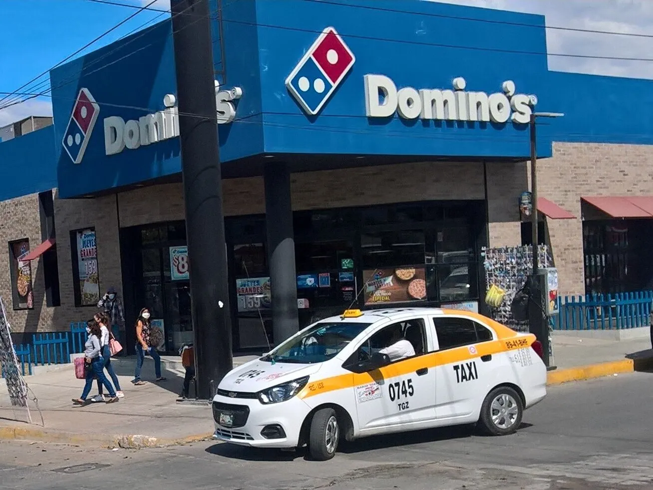 Sucursal de Domino's en Tuxtla Gutiérrez, Chiapas, con el logotipo y el nombre en el muro azul y un taxi estacionado al frente.