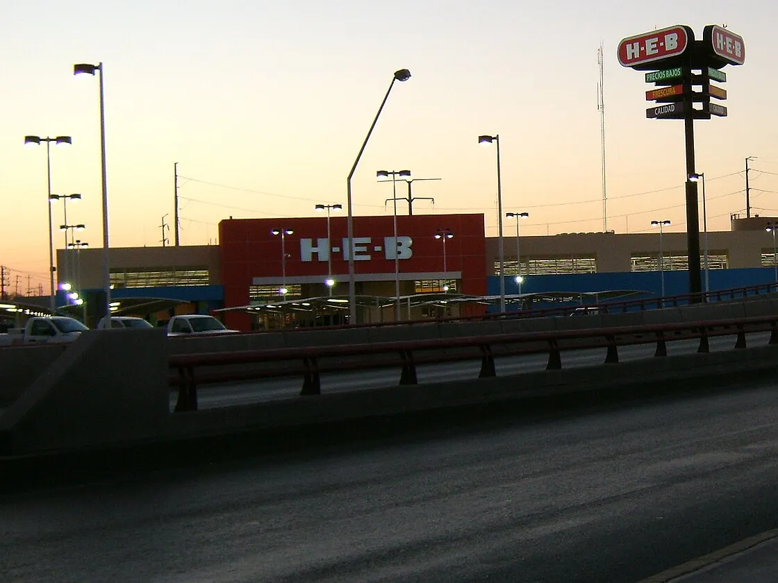 Tienda HEB de Piedras Negras, Coahuila, al atardecer, con el letrero rojo de H-E-B en la fachada y su anuncio alto junto al estacionamiento.