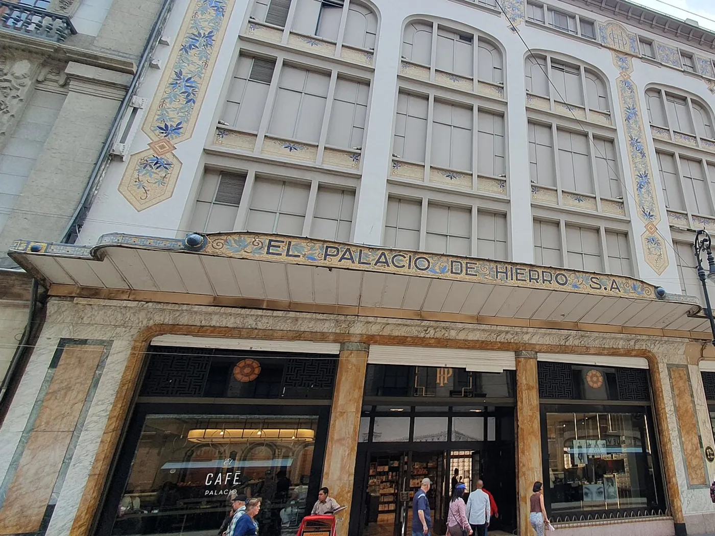 Entrada de El Palacio de Hierro en el Centro Histórico de la Ciudad de México, con la marquesina «EL PALACIO DE HIERRO, S.A.» sobre las puertas y el Café Palacio a la izquierda.