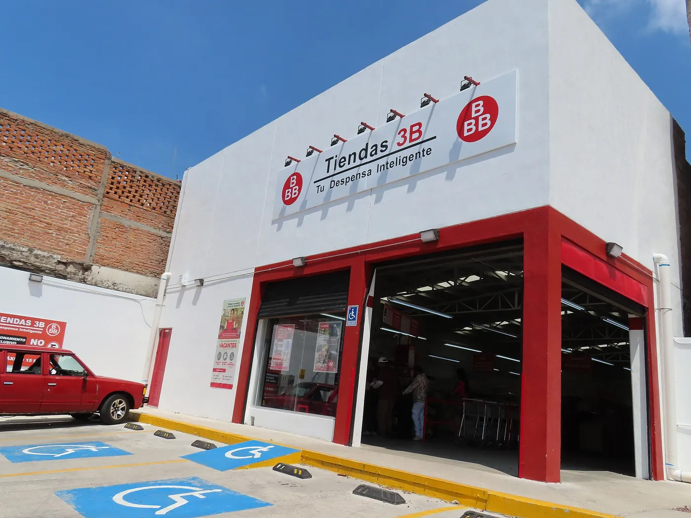 Fachada de una sucursal de Tiendas 3B en el centro de Aguascalientes, con el letrero «Tiendas 3B, Tu Despensa Inteligente», el marco rojo de la entrada y cajones de estacionamiento al frente.